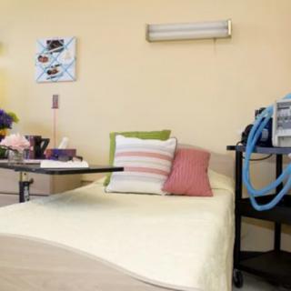 A tidy hospital room featuring a bed with colorful pillows, flowers on a side table, and medical equipment nearby.