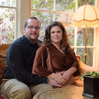 A couple sitting together on a sofa in a cozy living room with a large window and lamp.
