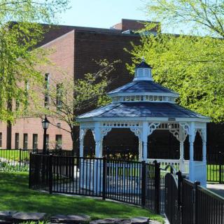 A white gazebo in a park setting with a brick building and trees in the background.