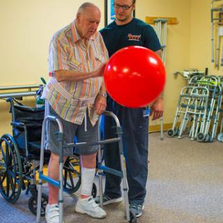 An elderly man receives assistance during a rehabilitation session, engaging with a red ball while using a walker.