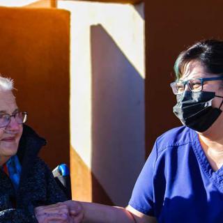 A healthcare worker wearing a mask interacts with an elderly woman outdoors.