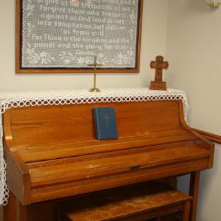 A wooden piano placed in a church setting, accompanied by a decorative cross and religious text.
