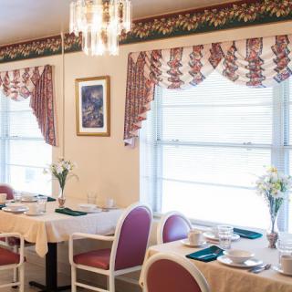 A cozy dining room with elegant decor, featuring tables set for a meal, intricate curtains, and a chandelier.