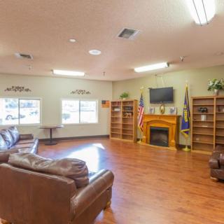 A spacious living room with comfortable seating, a fireplace, and a television mounted above. The room features bookshelves and flags, along with a wooden floor.