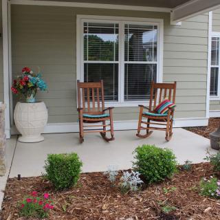 A cozy porch with two wooden rocking chairs and a decorative vase, surrounded by a small garden.