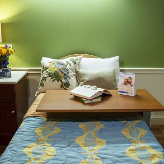 A cozy bedroom setup with a bed featuring decorative pillows, a bedside table with a lamp and flowers, and a book resting on a wooden tray.