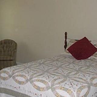 A cozy bedroom with a quilted bed, red pillow, and a wicker chair.