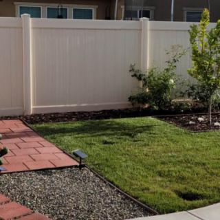 A backyard garden featuring a white fence, green lawn, and a brick pathway.