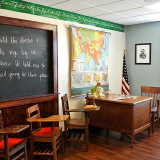 A vintage-style classroom with a chalkboard, wooden desk, and chairs, adorned with a world map and an American flag.
