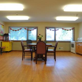 A well-lit dining room with a central table and chairs, featuring large windows and hardwood flooring.