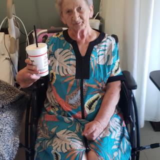 An elderly woman enjoying a drink while seated in her wheelchair, dressed in a vibrant floral-patterned dress.