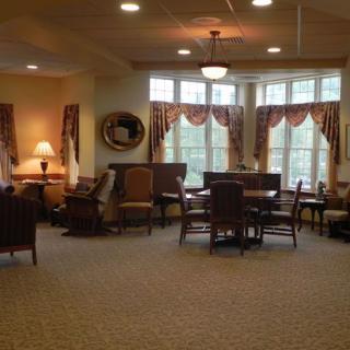 A cozy living room area with furniture, including tables and chairs, lamps, and large windows adorned with curtains.