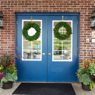 A pair of blue double doors with green wreaths, surrounded by plants, set against a brick wall.