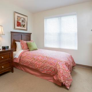 A cozy bedroom featuring a single bed with pink bedding, a nightstand with a lamp, and artwork on the wall.