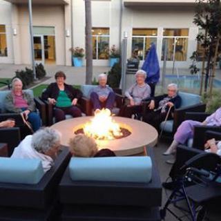 A group of seniors enjoys socializing around a fire pit in an outdoor seating area.
