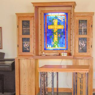 A chapel interior featuring a stained glass window with a cross, surrounded by wooden furniture.