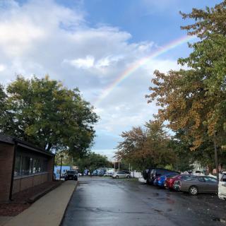 A street view with a rainbow arching over the trees on a cloudy day.