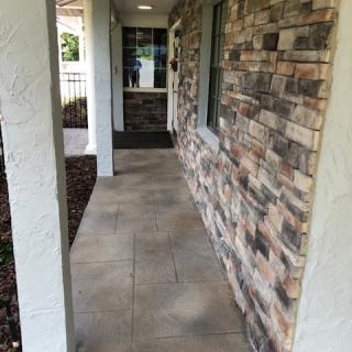 A stone-paved walkway with a brick wall and columns leading to a house entrance.