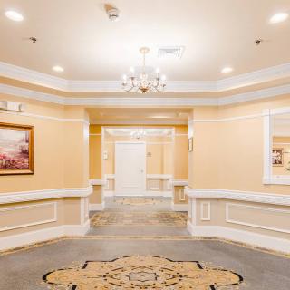 A well-lit hallway with elegant interior design features a chandelier, paintings, and a patterned carpet.