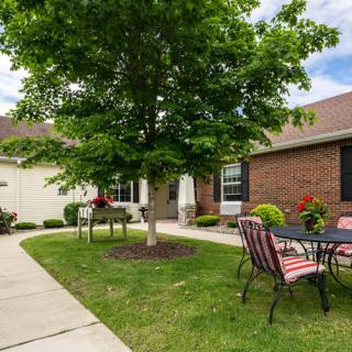 A cozy outdoor patio area with a table and chairs set under a tree, surrounded by a well-maintained lawn and brick house.