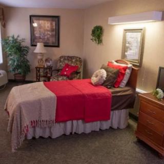 A cozy bedroom with a neatly made bed, accentuated with red and beige bedding, a lamp on the dresser, and a plant in the corner.