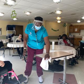 A staff member in personal protective equipment is cleaning a table in a nursing home facility.