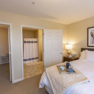 A cozy bedroom featuring a neatly made bed, adjacent nightstand with a lamp, and an open door leading to a bathroom.