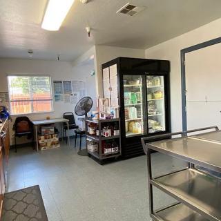 A small office space with a desk, refrigerator, shelving, and fan.