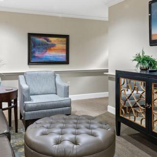 A cozy living room with a gray armchair, tufted ottoman, and framed artwork on the wall.