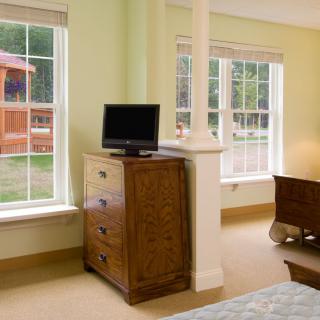 A cozy bedroom featuring a wooden dresser with a television, flanked by large windows letting in natural light.