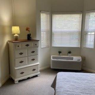 A cozy bedroom corner featuring a chest of drawers, a lamp and a large window with blinds allowing in daylight.
