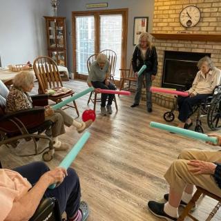 A group of seniors engaging in a seated exercise activity using pool noodles in a cozy living room setting.