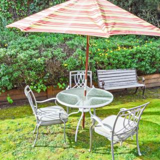 A garden scene featuring a patio set with a striped umbrella, surrounded by greenery.