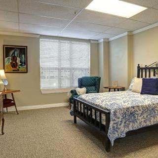 A cozy bedroom featuring a bed with floral bedding, an armchair by the window, and decorative elements including a lamp and artwork.