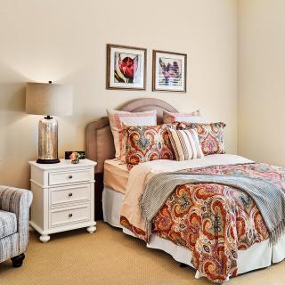 A cozy bedroom with patterned bedding, a nightstand with a lamp, and framed artwork on the wall.