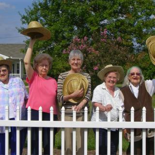 A group of women standing by a white fence, holding hats, enjoying a sunny day.