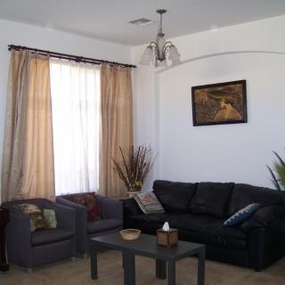 A cozy living room with a black sofa, armchair, and coffee table. Light filters through the curtains, highlighting the wall art and decorative elements.