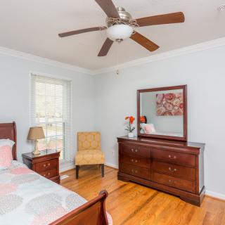 A bright bedroom featuring a ceiling fan, wooden dresser with a mirror, chair, and a lamp on hardwood floors.