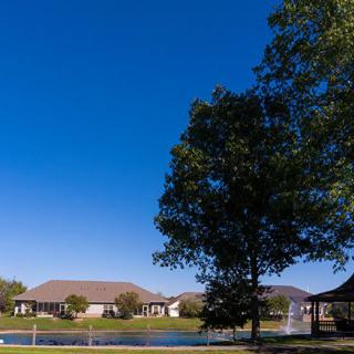 A serene landscape featuring a lake with a fountain, surrounded by trees and houses under a clear blue sky.