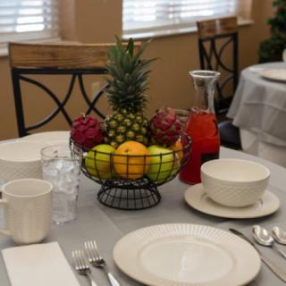 A neatly set dining table featuring a fruit basket centerpiece filled with an assortment of fruits.