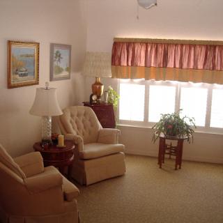 A cozy living room with armchairs, a lamp, and wall art, featuring soft natural light from a large window.