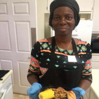 A woman in a kitchen wearing an apron and gloves, holding a plate of food.