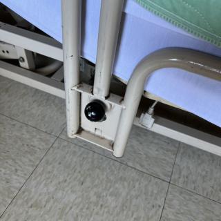 The image shows a close-up of a hospital bed's frame and wheel, positioned on a tiled floor.