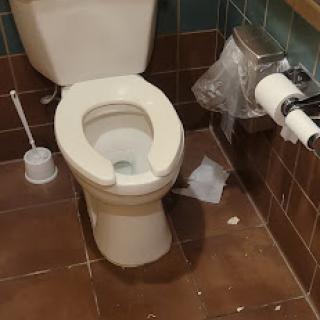 A view of a toilet in a restroom, showing cleaning supplies nearby.