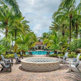 A tropical outdoor area featuring a fire pit surrounded by chairs with a pool in the background and lush palm trees.