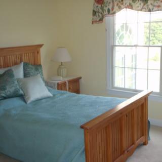 A cozy bedroom with a single bed, decorated with blue bedding and pillows, a bedside lamp, and a large window.