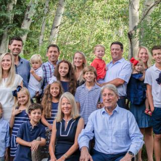 A large family gathers for a cheerful group photo among lush trees.