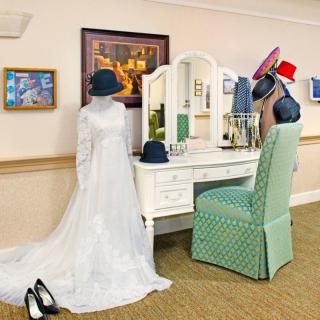 A mannequin in a vintage wedding dress stands by a vanity table adorned with various hats.