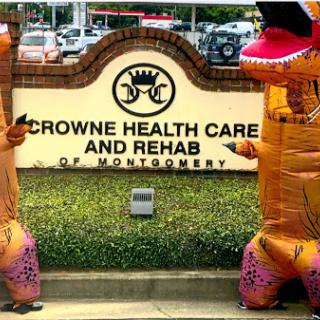 Two individuals in inflatable dinosaur costumes pose in front of Crowne Health Care and Rehab in Montgomery.
