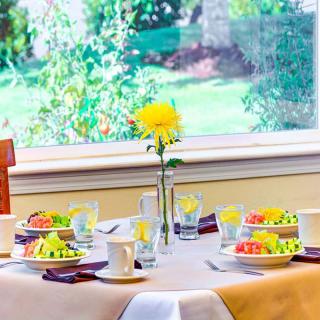 A beautifully set breakfast table with fresh fruit, cups, and a vase with a yellow flower by the window.